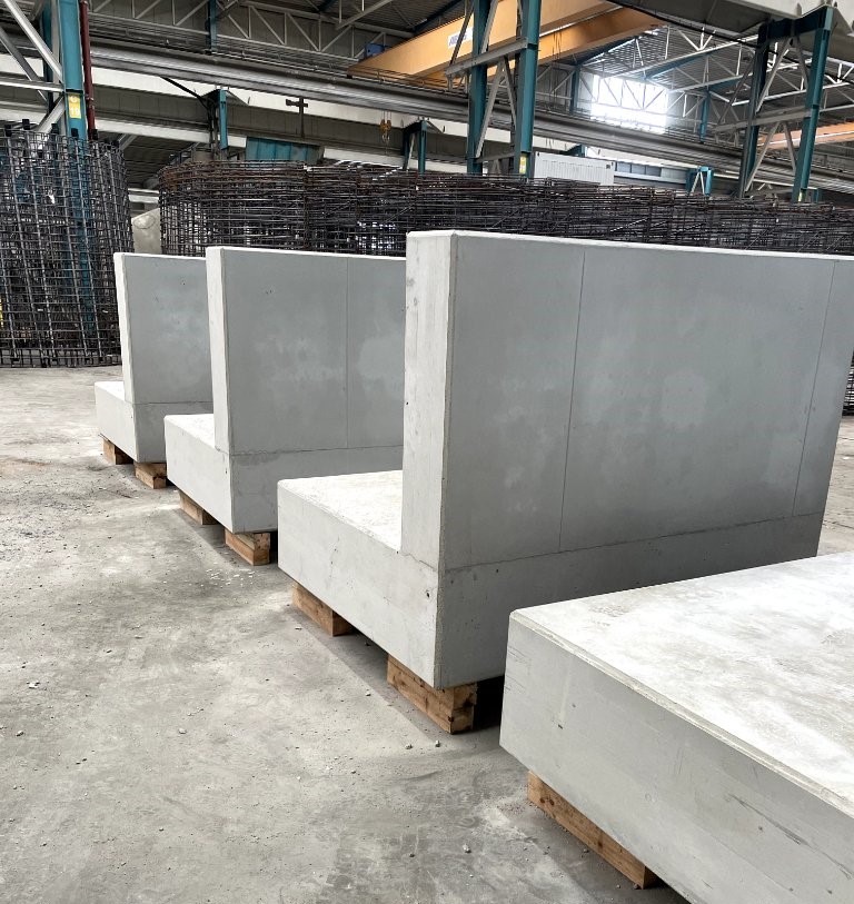 Foto von vier grauen Beton-Winkelstützelementen auf Holzpaletten in einer Industriehalle, mit Armierungseisen bereit für den weiteren Bau. Sie sind gestaffelt aufgestellt, was die Dimensionen und die stabile Bauweise betont.
